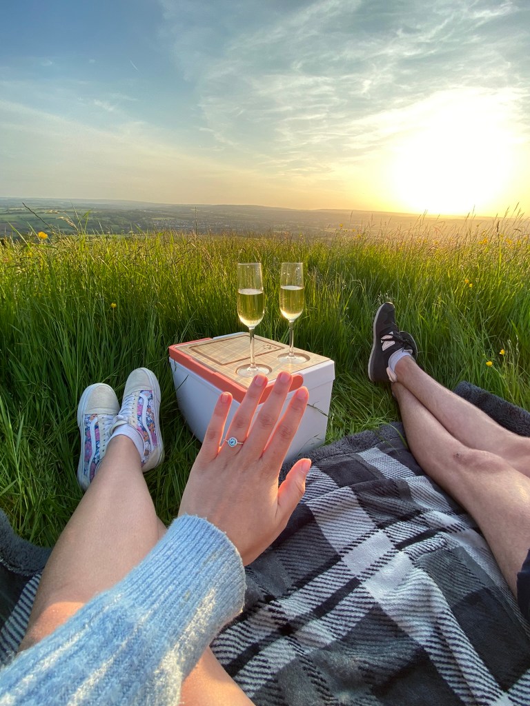 A snapshot from the happy couple's engagement. The author is showing her engagement ring on here finger with two glasses of champagne and a sunset in the background. The location of the picture is a field on Kelston Round Hill in Bath.